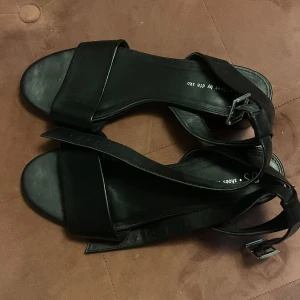 Svarta sandaler i skinn med spänne - Stilrena svarta sandaler från Shoes by Din Sko i mjukt skinn. De har bred rem över foten, öppen tå och justerbart spänne vid ankeln. Perfekta för en clean och minimalistisk look under varma dagar.