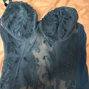 Blå korsett med broderade detaljer -  blå korsett med genomskinliga meshpartier och broderade blommönster. Kuporna är formade och har spetskant upptill. Axelbanden är justerbara och ryggen har klassisk hysk- och hakstängning. Perfekt för dig som vill ha något extra till outfiten.