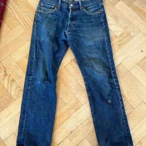 Levi's 501 blå jeans straight fit - Klassiska Levi's 501 jeans i blå denim med straight fit och fem fickor. Snyggt slitna detaljer och ikonisk läderpatch bak i midjan. Jeansen har orange sömmar och knappgylf. Perfekt för dig som gillar tidlös streetstil.
