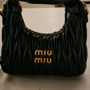 Svart quiltad handväska från Miu Miu - Snygg svart handväska från Miu Miu med quiltad design och guldiga detaljer. Väskan har ett mjukt, puffigt handtag och en praktisk dragkedja upptill. Materialet är skinn och loggan i guld ger en lyxig vibe. Perfekt storlek för det viktigaste.