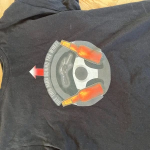 Svart t-shirt med ölflaskor från Gildan - Svart t-shirt från Gildan med tryck av två ölflaskor och en ratt på bröstet, samt texten 'Get this man behind the wheel'. Gjord i heavy cotton som känns rejäl och skön mot huden. Perfekt för dig som gillar roliga motiv och vill sticka ut.