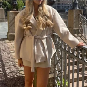Beige omlottjacka med bälte från Gina Tricot - Stilren beige omlottjacka från Gina Tricot med bälte i midjan och bred krage. Jackan har lång ärm och en loose passform som ger en avslappnad men trendig look. Perfekt för lager-på-lager under vår och höst.