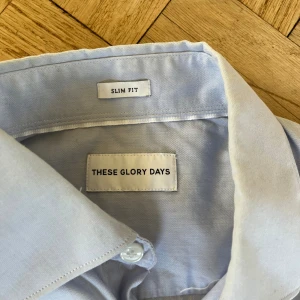 Ljusblå skjorta These Glory Days S - Snygg ljusblå skjorta från These Glory Days i slim fit-modell. Skjortan har klassisk krage, långa ärmar och vita knappar framtill. Tillverkad i mjuk bomull som känns skön mot huden. Perfekt för dig som gillar stilrena och enkla plagg.