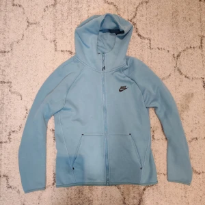 Ljusblå Tech Fleece från Nike - Säljer en ljusblå hoodie från Nike i storlek S. Tröjan har dragkedja framtill, två fickor och en skön huva. Nike-loggan är tryckt på bröstet. Perfekt för chill dagar eller när du vill ha en sportig look.