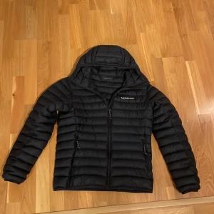 Svart dunjacka Peak Performance M - Svart dunjacka från Peak Performance med huva och snygga horisontella quiltade paneler. Jackan har dragkedja framtill och två sidofickor med dragkedja. Perfekt för kalla dagar och har en stilren look med diskret logga på bröstet.