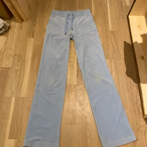 Juicy couture byxor  - Säljer ett par ljusblå juicy couture byxor med bred resår i midjan och snörning framtill. Byxorna har raka ben, stora fickor fram och bak med knappdetaljer. Materialet är mjukt och har en lätt glansig finish som ger en chill vibe. 