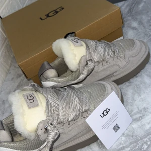 Beige sneakers från UGG med fårskinn - Snygga beige sneakers från UGG med breda snören och fluffigt vitt fårskinn på plösen. Ovandelen är i mocka och mesh, med en platt sula i gummi. UGG-logga på plösen och hälen. Perfekta för dig som vill ha både stil och komfort under höst och vinter.