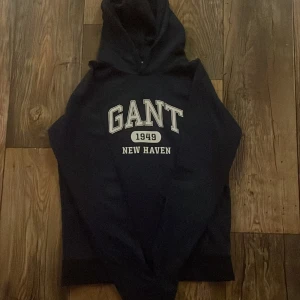 Mörkblå hoodie från GANT med tryck - Snygg mörkblå hoodie från GANT med stort vitt tryck på bröstet där det står 'GANT 1949 NEW HAVEN'. Klassisk huva och ribbade muddar. Perfekt för dig som gillar college-vibe och vill ha en bekväm tröja med coolt tryck. Pris går att diskutera.