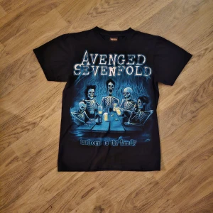 Avenged Sevenfold t-shirt Solid Rock - Svart t-shirt med Avenged Sevenfold-tryck fram och bak. Framsidan har ett stort motiv med skelett runt ett bord och texten 'welcome to the family'. Baksidan har bandets logga med dödskalle och fladdermusvingar. 
