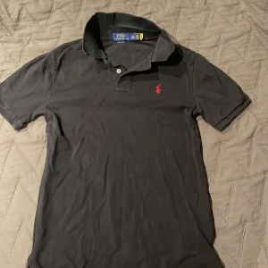 Svart pikétröja från Polo Ralph Lauren - Svart pikétröja från Polo Ralph Lauren med klassisk passform och korta ärmar. Tröjan har en snygg krage, två knappar vid halsen och den ikoniska röda Polo-loggan broderad på bröstet. Tillverkad i mjuk bomull som känns skön mot huden. Funkar även t storlek xs och s. Ny pris 1000