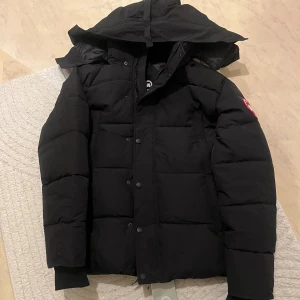 Canada goose jacka - Svart pufferjacka från Canada Goose med huva och klassisk logotyp på ärmen. Jackan har knappar och dragkedja framtill, quiltad design och ribbade muddar vid ärmslut. Perfekt för kalla dagar och har en clean, stilren look. OBS dragkedjan är lite svår men går att stänga 