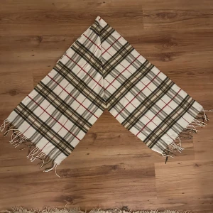 Rutig kashmirhalsduk från Burberrys - Sällsynt rutig halsduk från Burberrys i 100% kashmir. Färgerna är beige, svart, vit och röd med fransar i ändarna. Den är mjuk, lyxig och har den ikoniska Burberry-rutan som aldrig går ur tiden. Perfekt accessoar för att lyfta din outfit.
