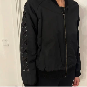 Svart bomberjacka med snörning - Svart bomberjacka med cool snörningsdetalj längs båda ärmarna och dragkedja framtill. Mörkblå. Storlek M/L men passar S/M. 