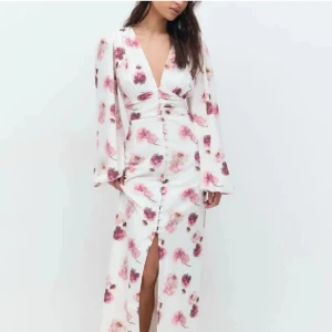Vit långklänning med rosa blommor - Supersnygg långklänning i vitt med blommigt mönster i rosa och lila toner. Klänningen har v-ringning, långa vida ärmar och knappar framtill med en slits. Figurnära passform i midjan och flowy kjol. Perfekt för sommarens alla tillfällen!