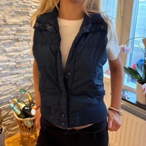 Mörkblå dunväst Abercrombie & Fitch - Snygg mörkblå dunväst från Abercrombie & Fitch med ribbad krage och nederkant. Västen har tryckknappar framtill, två snedställda fickor och quiltad design. Perfekt att slänga över en hoodie eller t-shirt för en chill look. Finns små fläckar