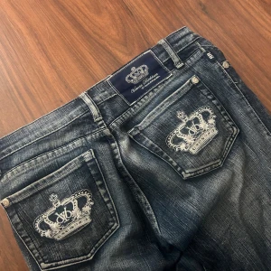 Victoria Beckham jeans med kronbrodyr - Snygga mörkblå jeans från Victoria Beckham med coola vita kronbroderier på bakfickorna. Klassisk femficksmodell med silverfärgade nitar och slitningar för en edgy look. Passar dig som vill sticka ut med exklusiva detaljer.