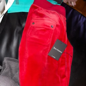 Röda mjukisbyxor Juicy Couture - Snygga röda mjukisbyxor från Juicy Couture med bred resår i midjan och en stor ficka med knappar på sidan. Byxorna har en mjuk och glansig yta som känns lyxig och skön mot huden. Perfekta för chill eller häng med kompisar.