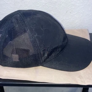 Svart Gucci keps med mesh - Snygg svart keps från Gucci med klassiskt GG-mönster på framsidan och mesh på sidorna och bak. Kepsen har böjd skärm och justerbar rem baktill. Märkeslapp i skinn bak. Perfekt för en trendig streetstyle-look.