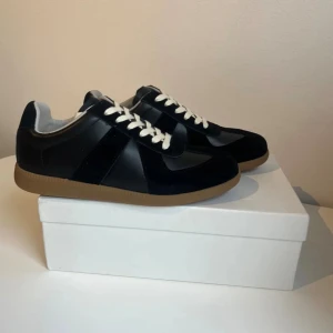 Svarta Maison margiela skor  - Maison Margiela sneakers i svart skinn och mocka med vita snören och brun gummisula. Klassisk low-top siluett med rund tå och stilren design. Perfekta för dig som gillar minimalistisk och tidlös stil. Äkta 