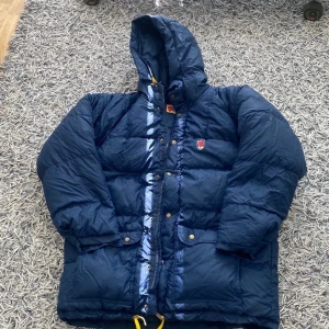 Mörkblå dunjacka från Fjällräven - Säljer en mörkblå dunjacka från Fjällräven med huva och klassisk logga på bröstet. Jackan har stora fickor med knappar, dragkedja och tryckknappar framtill samt gul detalj nertill. Perfekt för kalla dagar. Jackan är också inte så använd och i väldigt bra skick!