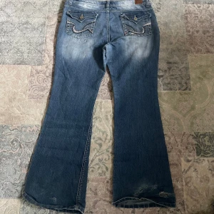 Blå flare jeans från l.e.i, strl 13 - Snygga blå jeans från l.e.i i modellen Sophia Hipster Flare. Jeansen har låg midja, utsvängda ben och coola bakfickor med broderade detaljer och knappar. Tillverkade i klassiskt denimtyg med slitningar och ljusare partier framtill och baktill. Lite defekter på slutet av benen (kolla sista bilderna)