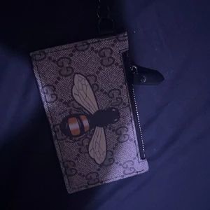 Gucci plånbok med bi och axelrem - Snygg plånbok från Gucci med klassiskt GG-mönster och stor bi på framsidan. Utrustad med svart läder på baksidan, kortfack och dragkedja. Avtagbar axelrem i grönt och rött för extra stilpoäng. Perfekt för dig som vill ha något unikt och trendigt. ÄKTA  pris kan diskuteras vid snabbaffär