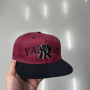  Yankees keps med svart skärm - Snygg bordeaux keps med svart skärm och broderad Yankees-logga i svart och vitt framtill. Klassisk snapback-modell med platt skärm och grönt underskärm. Perfekt för dig som vill ha streetstyle med sportig vibe.