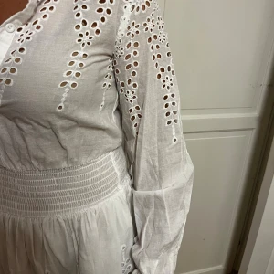 Vit broderad kortklänning Loavies  - Superfin vit kortklänning från Loavies i storlek XS. Klänningen har broderade blommor och hålmönster, smock i midjan och knappar framtill. Långärmad med lätt puff och spetsdetaljer vid fållen. Perfekt för sommarens alla tillfällen!