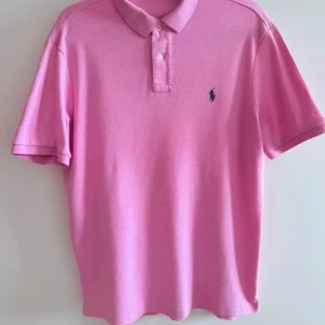 Rosa pikétröja från Polo Ralph Lauren - Snygg rosa pikétröja från Polo Ralph Lauren med klassisk krage, korta ärmar och knappar framtill. Ikoniska loggan broderad på bröstet. Perfekt för dig som vill ha en stilren och fräsch look.