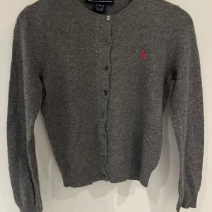 Grå kofta från Ralph Lauren Sport - Mysig grå kofta från Ralph Lauren Sport med rund halsringning och knappar framtill. Liten rosa logga broderad på bröstet. Tillverkad i en lyxig mix av ull, nylon, viskos och kashmir. Perfekt för lager-på-lager under kyliga dagar.                                  OBS passar xs det är M i Amerikansk ungdoms stl