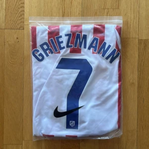 Atletico Madrid Griezmann tröja Nike M - Säljer en officiell Atletico Madrid fotbollströja med Griezmann och nummer 7 på ryggen. Tröjan är kortärmad, har rödvita vertikala ränder, blå Nike-logga och klubbmärke på bröstet. Materialet är Dri-Fit polyester och har sponsortryck från Riyadh Air.