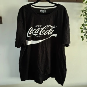 Svart Coca-Cola t-shirt XXL - Svart t-shirt från Coca-Cola i storlek XXL med klassiskt vitt tryck framtill. Oversized passform och rund halsringning. Perfekt för dig som gillar streetstyle och vill ha en chill vibe. Gjord i mjuk bomull för extra komfort. Använt skick, sitter som XL