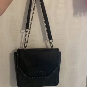 Stella McCartney väska - Snygg svart axelväska från Stella McCartney med kedjedetaljer längs kanten och axelrem i både kedja och skinn. Väskan har lock med logotyp framtill och ett rymligt innerfack med mönstrat foder. Man kan ändra om man vill ha en lång eller kort kedja. Använd 1-2 gånger och inga defekter💕skriv för fler bilder, pris kan diskuteras💗är inte 100% säker på att denna är riktig då jag fick den i present och inte vet var den är ifrån därav priset💕💗