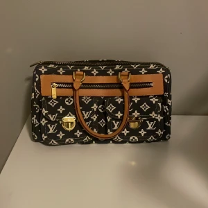 svart handväska med monogrammönster - Snygg svart handväska med klassiskt vitt monogrammönster. Väskan har bruna skinnhandtag, guldiga metalldetaljer och flera fack framtill med dragkedja och spännen. Perfekt storlek för att få plats med allt du behöver. Tyvärr små skador som man ser på bild 4-5. Kan gå ner i pris vid snabb affär