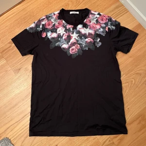 Svart blommig t-shirt från Givenchy - Svart t-shirt från Givenchy med korta ärmar och rund hals. Framsidan och baksidan har tryck av rosa och röda blommor samt silverfärgade stjärnor över axlar och bröst. Tillverkad i mjuk bomull för en skön känsla och snygg passform. Storlek S sitter som M