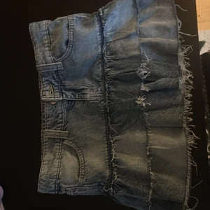 Blå jeanskjol med volang och råa kanter - Säljer en blå jeanskjol med volangdetaljer och råa, fransiga kanter. Kjolen har klassiska fickor fram och hällor för bälte. Perfekt för dig som gillar en edgy och trendig stil.