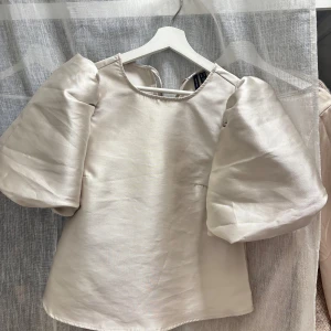 Champagnefärgad blus med puffärm Vero Moda - Säljer en champagnefärgad blus från Vero Moda med stora puffärmar och rund halsringning. Blusen har en snygg knytning i nacken och är tillverkad i ett glansigt material som ger en lyxig känsla. Passar perfekt till en utekväll me tjejerna!!💕💕