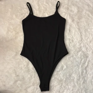 Svart enkel baddräkt med smala axelband - Snygg svart boody med smala axelband och klassisk, stilren design. Hög benskärning och rundad halsringning ger en modern look. Perfekt för fest eller dejt. Materialet är stretchigt och bekvämt.