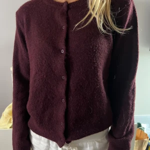 Mörklila stickad kofta med knappar - Mysig mörklila stickad kofta med rund halsringning och knäppning framtill. Koftan har långa ärmar och en lite fluffig känsla i materialet. 