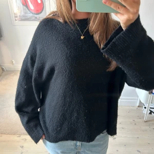 Svart stickad tröja från Gina Tricot - Mysig svart stickad tröja från Gina Tricot med avslappnad passform och bred ribbad krage. Tröjan har långa ärmar och är perfekt för lager-på-lager. Enkel och stilren design som funkar till allt.