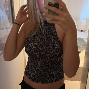 Leopardmönstrad halterneck topp - Säljer en snygg croppad halterneck topp med leopardmönster i svart och grått som är den perfekta fest toppen. Toppen är ärmlös och har en tight passform som framhäver axlarna. Perfekt för dig som vill sticka ut med ett djurmönster och gillar trendiga plagg.