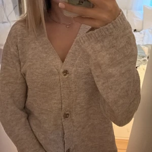 Beige stickad kofta med knappar - Mysig beige stickad kofta med v-ringning och stora knappar framtill. Koftan har långärmade ärmar och en relaxed passform, perfekt att slänga på sig när det är kyligt. Enkel och stilren design som passar till det mesta.