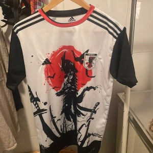Japan Adidas fotbollströja samurai. - Japan fotbollströja från Adidas med unikt samurai-tryck i svart och rött på både fram- och baksida. Tröjan är vit med svarta ärmar, röda detaljer vid kragen och har Japan JFA-emblem på bröstet. Tillverkad i lätt och ventilerande material, perfekt för match eller träning.