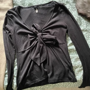 Svart långärmad topp med knyt Gina Tricot - Svart långärmad topp från Gina Tricot med snygg v-ringning och stor knytning framtill. Toppens material är mjukt och stretchigt, perfekt för att skapa en cool och trendig look. Passar dig som gillar stilrena plagg med en twist.