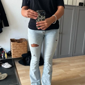 Ljusblå bootcut jeans med slitningar - Säljer ett par ljusblå jeans med bootcut passform och snygga slitningar på låret och knät. Jeansen har klassisk femficksdesign och är tillverkade i denim. Perfekta för dig som gillar en avslappnad och trendig stil.