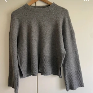 Grå stickad tröja med slits - Mysig grå stickad tröja med rund halsringning och långa ärmar. Tröjan har ribbade muddar och nederkant samt små slitsar på sidorna 