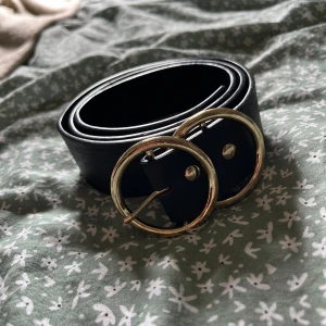 Svart bälte med rund guldfärgad spänne - Stilrent svart bälte i skinn med ett stort, runt guldfärgat spänne. Perfekt accessoar för att lyfta vilken outfit som helst med en modern touch. Bältet är brett och har en enkel men snygg design som passar till jeans eller klänning.