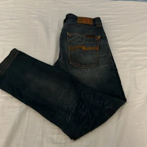 Mörkblå jeans från Nudie Jeans - Snygga mörkblå jeans från Nudie Jeans med klassisk orange kontrastsöm och ikonisk broderad bakficka. Jeansen har rak passform och är tillverkade i slitstarkt denim. Perfekta för dig som gillar en stilren och tidlös look. Det står att storleken är 29 34 men dem är uppsydda så dem är typ 32