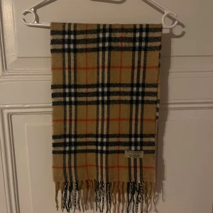 Rutig halsduk från Burberry i ull - Klassisk halsduk från Burberry i 100% lammull med ikoniskt rutigt mönster i beige, svart, vitt och röda detaljer. Mjukt material och fransar i ändarna. Perfekt accessoar för att lyfta din outfit.