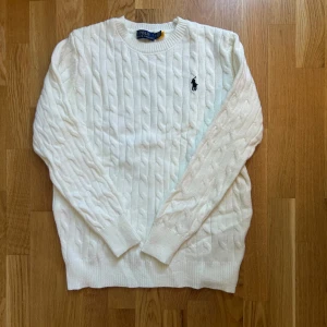 Vit kabelstickad tröja Polo Ralph Lauren - Kabelstickad vit tröja från Polo Ralph Lauren med klassisk rund halsringning och broderad marinblå logga på bröstet. Tröjan är långärmad och har ribbade muddar vid ärmslut och nederkant. Perfekt för lager-på-lager under höst och vinter.